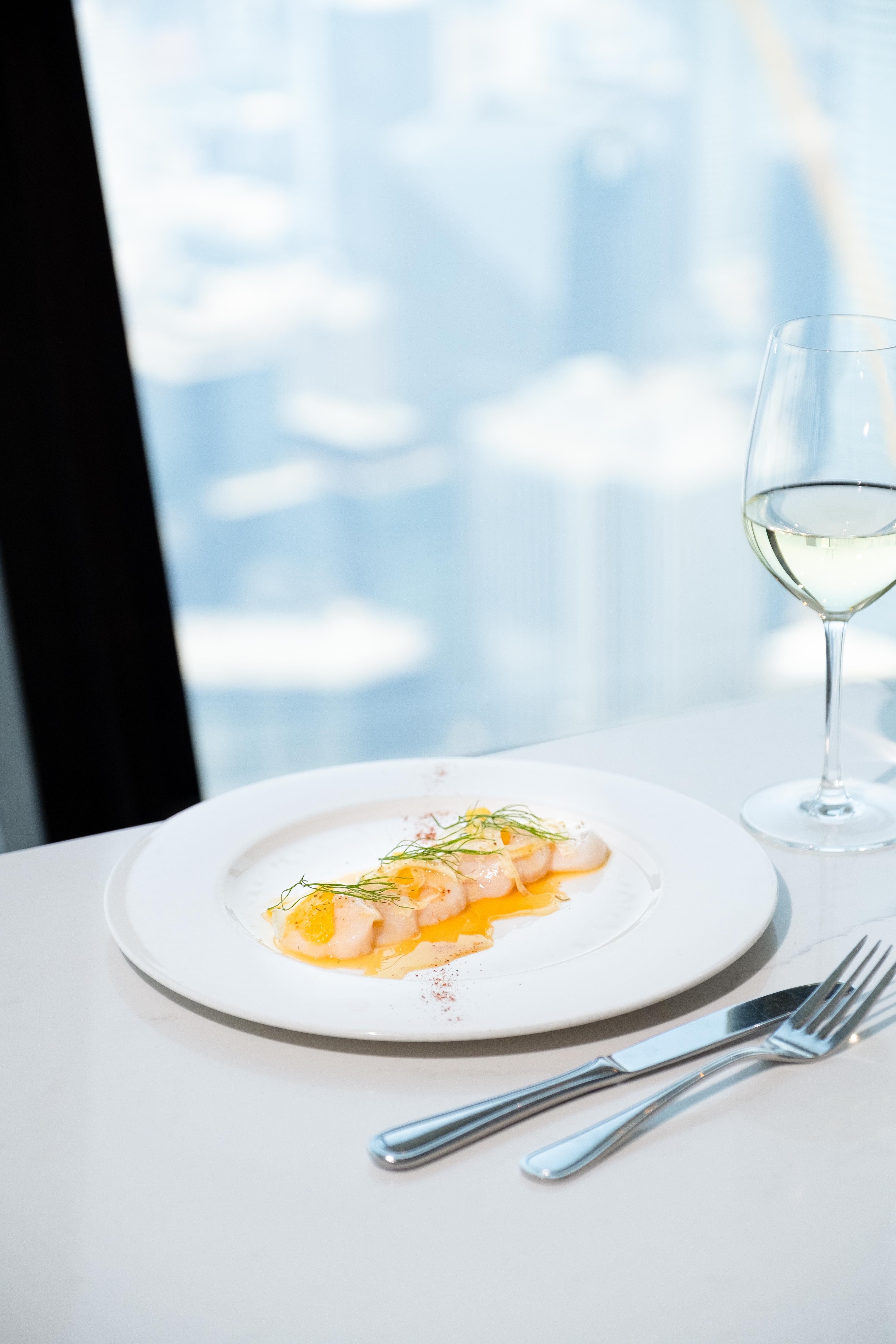 View of the scallops crudo on a white plate on a table at 360 with a glass of white wine and the city in the background.