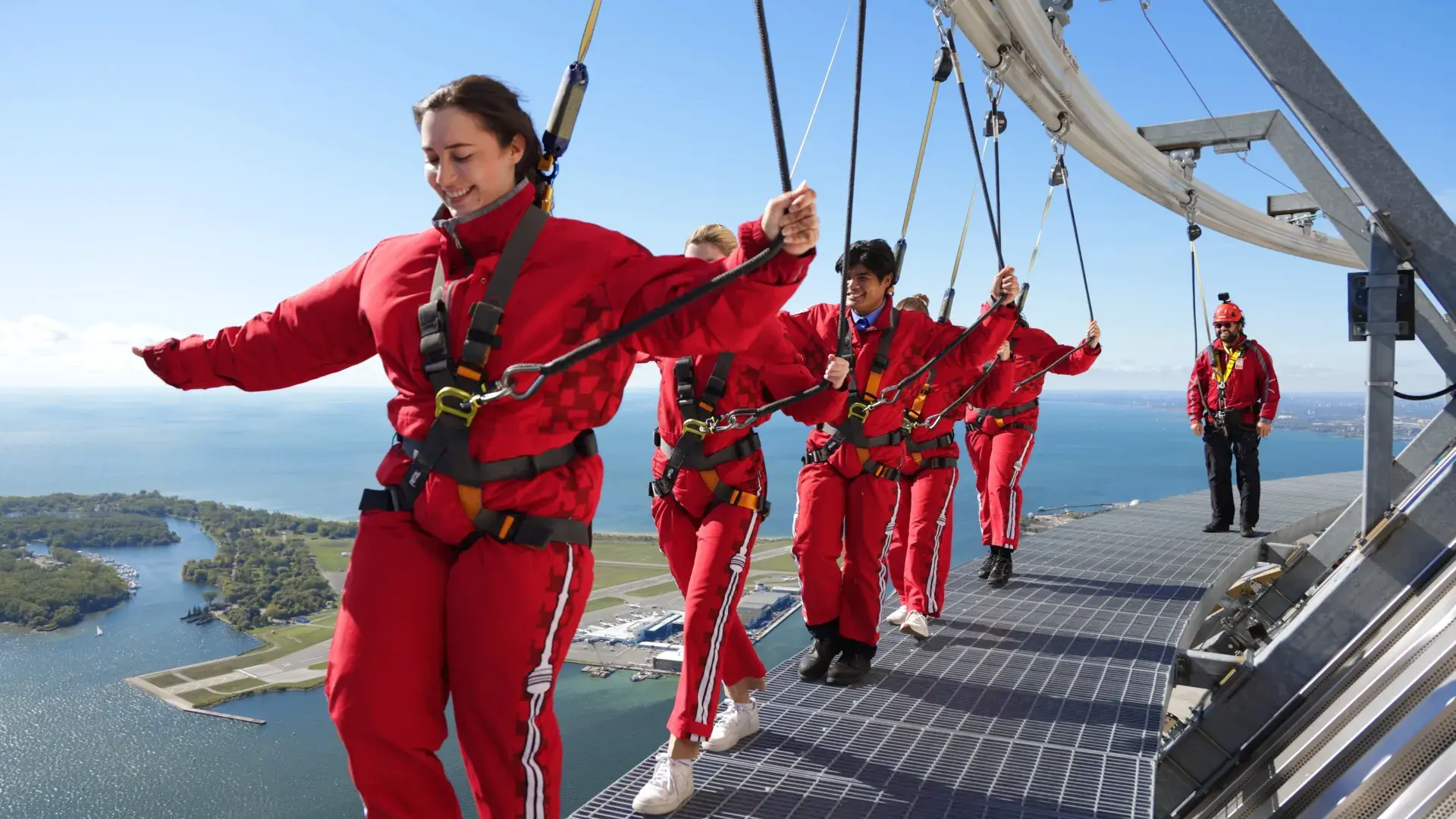 Group of EdgeWalk participants in red suits and safety harnesses walking along the platform, attached to overhead rails, with Lake Ontario and Toronto Islands visible below.