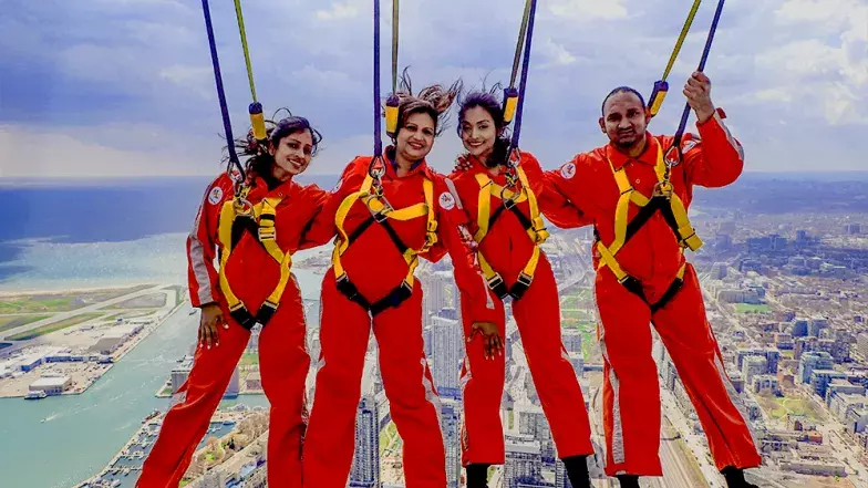 Four people, side by side leaning back over the edge. Far below and behind them is the city of Toronto on a clear summer day.