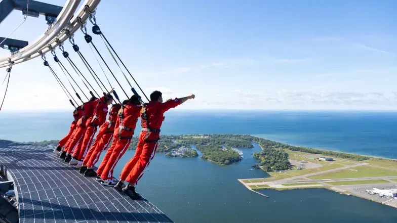 EdgeWalk participants lean outward from the platform while secured to overhead cables, facing Lake Ontario and the Toronto Islands.