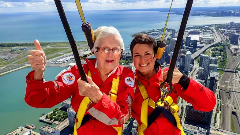 Two people with thumbs up and smiling with the city of Toronto far below on a bright but cloudy summer day.