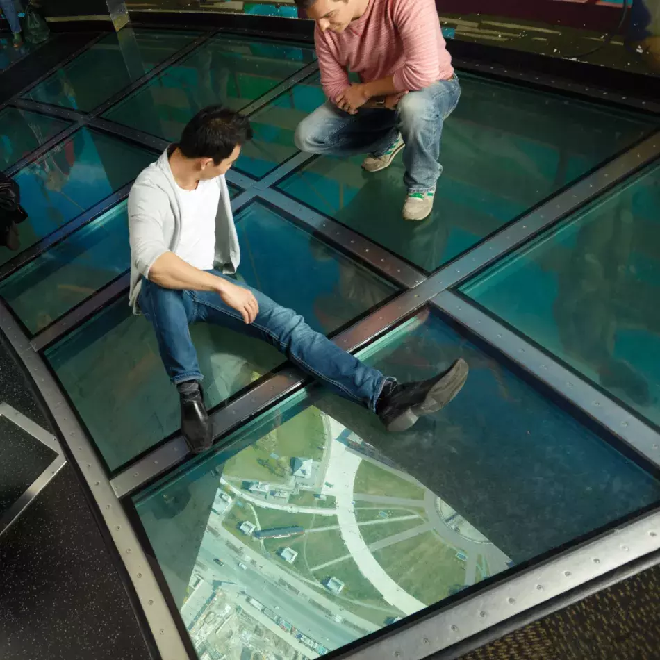 Two people sitting on the glass floor with the ground visible far below.