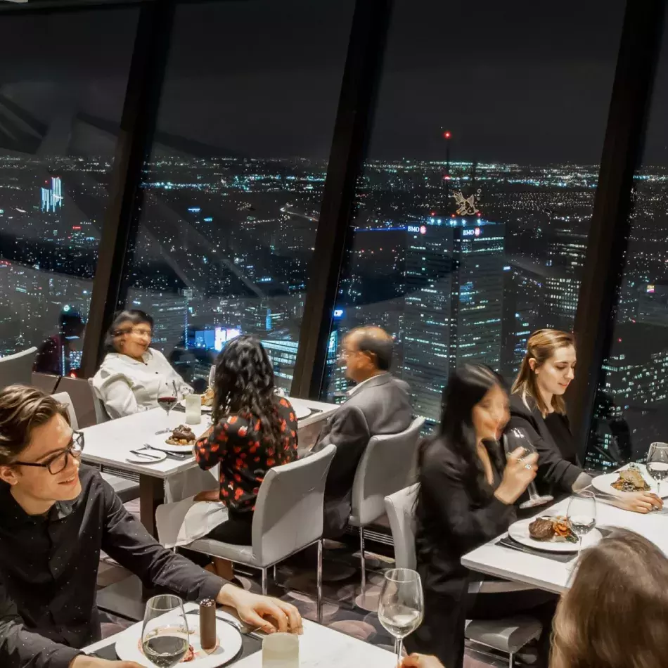 Six people sitting at three tables eating dinner with Toronto city lights at night in the background.