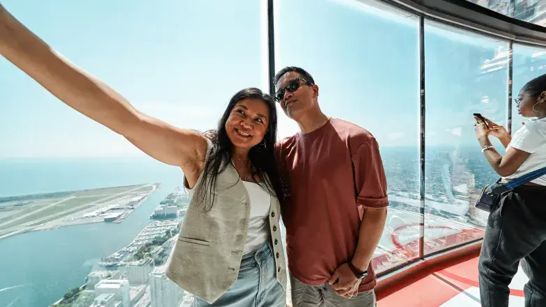 A couple taking a selfie at the CN Tower near a large window with a view of Lake Ontario and the city below.